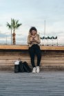 Задоволена кучерява молода жінка в повсякденному одязі посміхається, використовуючи мобільний телефон на лавці з сумками на сучасній набережній — стокове фото