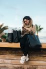 Задоволена кучерява молода жінка в повсякденному одязі посміхається, використовуючи мобільний телефон на лавці з сумками на сучасній набережній — стокове фото