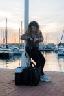 Вид збоку на стильну молоду жінку в повсякденному вбранні, що спирається на пост під час перевірки рюкзака на сучасному пірсі після покупки — стокове фото