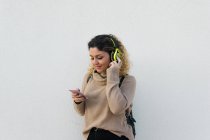 Молодая счастливая женщина в бежевом свитере улыбается, используя наушники и мобильный телефон с белой стеной на заднем плане — стоковое фото