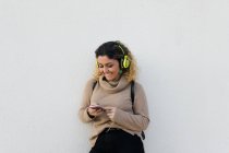 Jeune femme heureuse en pull décontracté beige souriant tout en utilisant un casque et un téléphone portable avec mur blanc sur fond — Photo de stock