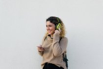 Молодая счастливая женщина в бежевом свитере улыбается, используя наушники и мобильный телефон с белой стеной на заднем плане — стоковое фото