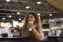 Dama contemporánea en suéter beige casual apoyando los codos en la mesa con café para ir en el aeropuerto sobre fondo borroso - foto de stock