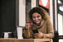 Mulher bonita feliz com cartão de crédito vermelho segurando telefone celular na mesa de madeira — Fotografia de Stock