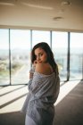 Back view of dreamy sensual beautiful woman in striped shirt showing naked shoulder while standing in room with panoramic windows looking ta camera — Stock Photo