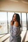 Back view of dreamy sensual beautiful woman in striped shirt showing naked shoulder while standing in room with panoramic windows looking ta camera — Stock Photo