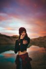 Romantische, jugendlich sensible, süße Frau in lässiger Kleidung, die wegschaut, während sie am Seeufer im bunten Sonnenuntergang steht — Stockfoto