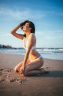 Vista lateral de alegre jovem turista em trajes de banho sentado de joelhos em luz solar na praia de areia durante as férias de verão para o mar — Fotografia de Stock