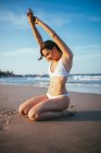 Seitenansicht einer fröhlichen jungen Touristin in Badebekleidung, die auf Knien im Sonnenlicht am Sandstrand während des Sommerurlaubs am Meer sitzt — Stockfoto