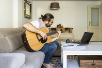 Vista laterale del musicista maschio creativo barbuto in abbigliamento casual seduto sul divano e suonare la chitarra acustica nel soggiorno moderno — Foto stock