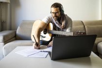 Músico barbudo en anteojos sentado en un sofá con guitarra y escribiendo notas en la mesa mientras usa auriculares y laptop - foto de stock