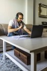 Ernsthafter bärtiger Mann in Freizeitkleidung und Brille schaut Laptop, während er im stilvollen Wohnzimmer Musik mit Kopfhörern hört — Stockfoto
