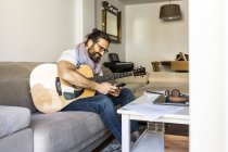 Uomo allegro con smartphone controllo chitarra sul divano — Foto stock