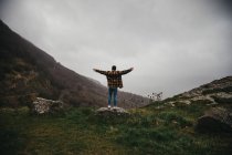 From below back view of unrecognizable male hiker in casual clothes standing on cliff and spreading arms in cloudy day in countryside — Stock Photo