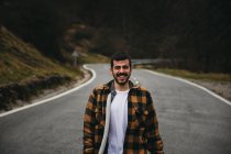 Bearded black haired cheerful man in casual wear smiling and looking at camera while standing on empty road in autumn countryside — Stock Photo