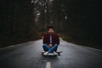 Беззаботный этнический парень в стильной повседневной одежде сидит со скрещенными ногами на скейтборде по асфальтовой дороге и смотрит в камеру среди осеннего леса — стоковое фото