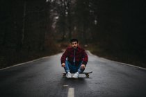 Беззаботный человек в стильной повседневной одежде сидит со скрещенными ногами на скейтборде по асфальтовой дороге и смотрит в камеру среди осеннего леса — стоковое фото