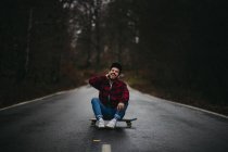 Безтурботний веселий чоловік у стильному повсякденному одязі сидить з схрещеними ногами на скейтборді на асфальтовій дорозі і дивиться на камеру серед осіннього лісу — стокове фото