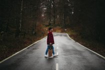 Vista lateral do jovem homem elegante em desgaste casual andando na estrada de asfalto com skate na mão no dia de outono — Fotografia de Stock