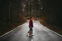 Вид сбоку на молодого стильного мужчину в повседневной одежде, идущего по асфальтовой дороге со скейтбордом в руке в осенний день — стоковое фото