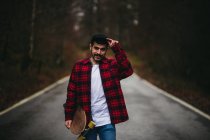 Jovem homem elegante em desgaste casual andando na estrada de asfalto com skate na mão no dia de outono olhando para a câmera — Fotografia de Stock