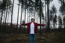 Jeune touriste barbu en tenue décontractée écartant les bras et levant les yeux au milieu de la forêt de conifères à feuilles persistantes sur ciel couvert — Photo de stock