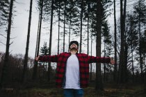 Young bearded male tourist in casual wear spreading arms and looking up among evergreen coniferous forest on overcast — Stock Photo