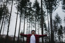 Joven turista barbudo en ropa casual extendiendo los brazos y mirando hacia arriba entre el bosque de coníferas de hoja perenne en el nublado - foto de stock