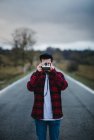 Homem irreconhecível em roupa casual elegante segurando câmera de fotos na frente do rosto enquanto está de pé na estrada no campo — Fotografia de Stock