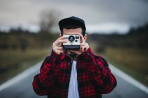 Unrecognizable man in stylish casual outfit holding photo camera in front of face while standing on road in countryside — Stock Photo