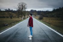 Side view of man in casual wear walking on empty asphalt road among green fields with cloudy sky on background — Stock Photo