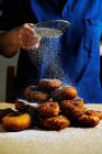 Persona irriconoscibile che utilizza il setaccio per versare zucchero a velo su una pila di ciambelle fresche durante la cottura della pasta in cucina — Foto stock