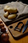 Vista superior de batatas cruas na mesa com azeite em ovos jar faca de cebola e descascador em tábua de corte de madeira na cozinha — Fotografia de Stock