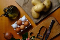 Vista dall'alto delle patate crude in tavola con olio d'oliva in vasetto uova coltellino cipolla e pelapatate su tagliere in legno in cucina — Foto stock