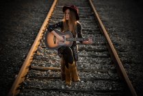 Full body stylish female playing guitar and looking away while sitting on rails during trip — Stock Photo