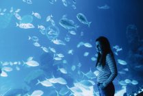 Vue latérale de joyeuse jeune femme aux cheveux noirs en tenue décontractée souriante et regardant vers le haut avec un aquarium moderne lumineux sur fond — Photo de stock