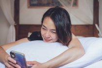 Веселая этническая женщина улыбается и просматривает смартфон, лежа на подушке в удобной постели дома — стоковое фото