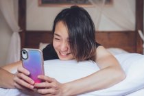 Весела етнічна жінка посміхається і переглядає смартфон, лежачи на подушці в зручному ліжку вдома — стокове фото