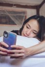 Dal soffietto allegro etnico femminile sorridente e smartphone di navigazione mentre sdraiato sul cuscino in comodo letto a casa — Foto stock