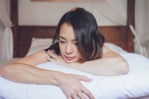 Optimista mujer asiática reflexiva mirando hacia otro lado acostado en la cama cómoda en la mañana en casa - foto de stock