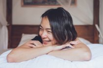Otimista mulher asiática olhando afastado deitado na cama confortável e rindo enquanto deitado na cama de manhã em casa — Fotografia de Stock