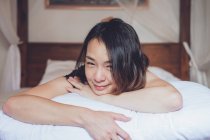 Optimistische Asiatin blickt in die Kamera, die auf einem bequemen Bett liegt und lacht, während sie morgens zu Hause auf dem Bett liegt — Stockfoto