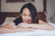 Оптимістична азіатка дивиться на камеру лежачу на зручному ліжку і сміється, лежачи на ліжку вранці вдома. — стокове фото