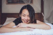 Оптимистичная азиатская женщина смотрит в камеру, лежащую на удобной кровати и смеется, лежа на кровати утром дома — стоковое фото