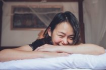 Optimistische Asiatin blickt in die Kamera, die auf einem bequemen Bett liegt und lacht, während sie morgens zu Hause auf dem Bett liegt — Stockfoto