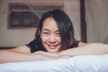 Mujer asiática optimista mirando la cámara tumbada en la cama cómoda y riendo mientras está acostada en la cama por la mañana en casa - foto de stock