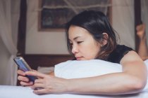 Allegro smartphone etnico femminile sorridente e di navigazione mentre sdraiato sul cuscino in un comodo letto a casa — Foto stock