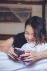 From bellow cheerful ethnic female smiling and browsing smartphone while lying on pillow in comfortable bed at home — Stock Photo