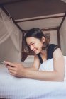 From bellow cheerful ethnic female smiling and browsing smartphone while lying on pillow in comfortable bed at home — Stock Photo