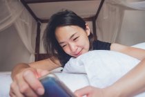 Від весела етнічна жінка посміхається і переглядає смартфон, лежачи на подушці в зручному ліжку вдома — стокове фото